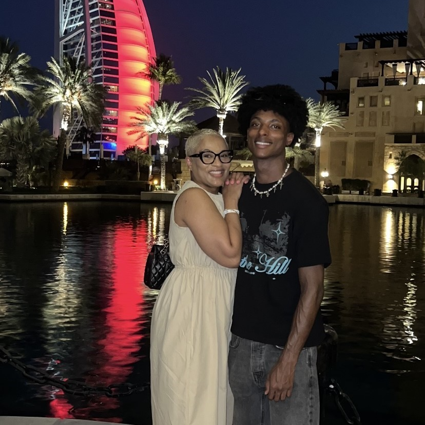 Photo of mother and grown up son standing next to each other, both smiling - beautiful exotic background, night time, with lit skyscrapers, also palm trees in area