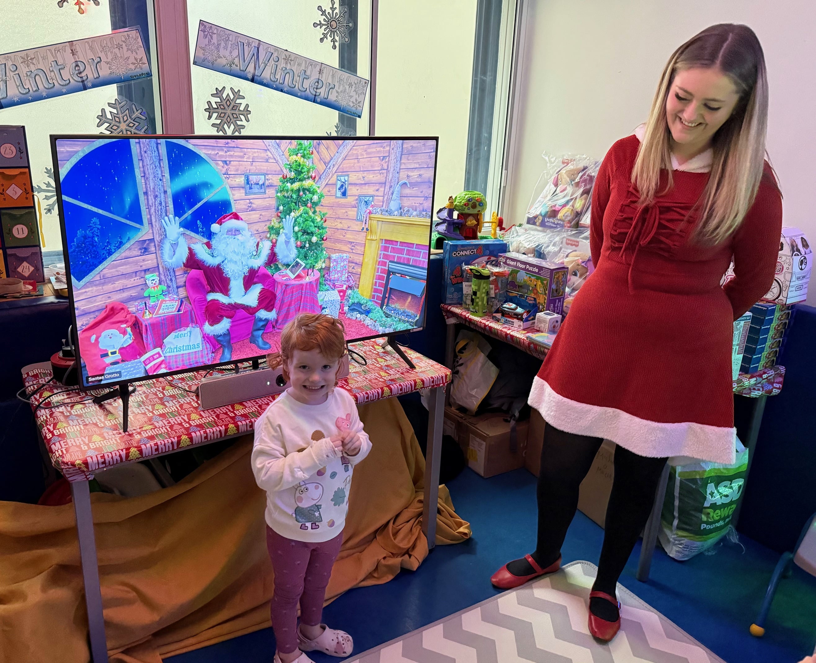 Staff member dressed as Santa helper, next to a smiling child, Santa on TV in background