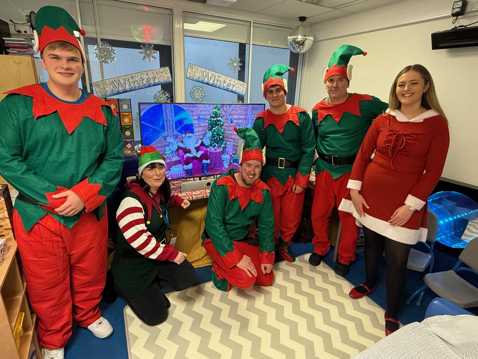 People dressed as elves, next to a TV gift, smiling, in a hospital setting