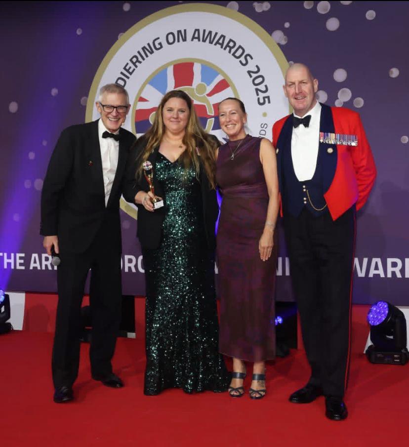 Lt Col Rachel Diss smiling, while holding her award at the ceremony's podium, with a few other people alongside her